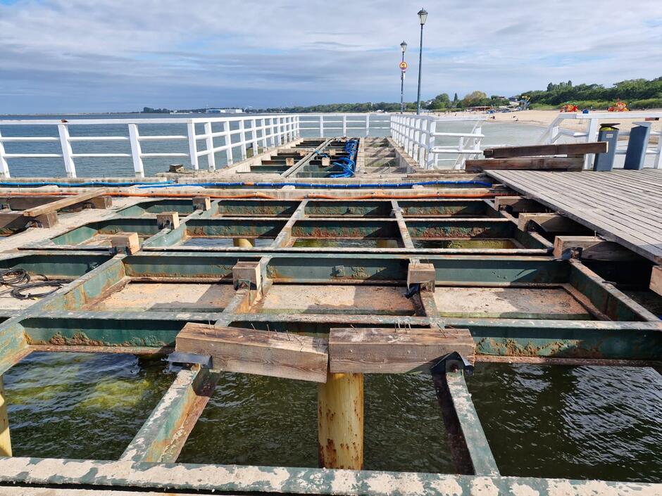 Na zdjęciu widać fragment molo w trakcie remontu, z widoczną stalową konstrukcją nośną nad wodą. W tle rozciąga się plaża oraz morze, a część pomostu wciąż posiada drewnianą nawierzchnię. 