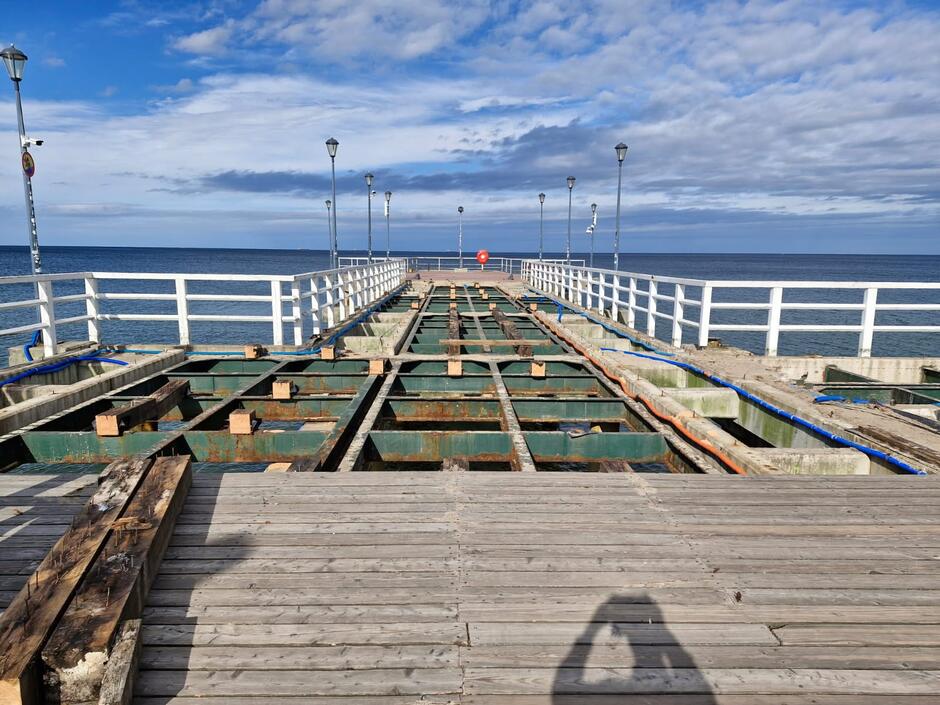 Na zdjęciu przedstawiono molo nad morzem w trakcie remontu, z widoczną odsłoniętą stalową konstrukcją i instalacjami. W tle rozciąga się spokojne morze i niebo z lekkim zachmurzeniem, a całość otacza biała balustrada z lampami. 