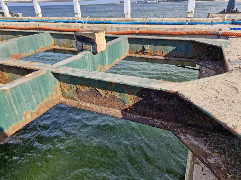 Na zdjęciu widoczna jest stalowa konstrukcja molo, silnie skorodowana i pokryta śladami rdzy w miejscach styku z betonem. Poniżej znajduje się woda morska, a w tle widać balustradę oraz fragmenty instalacji biegnących wzdłuż pomostu. 