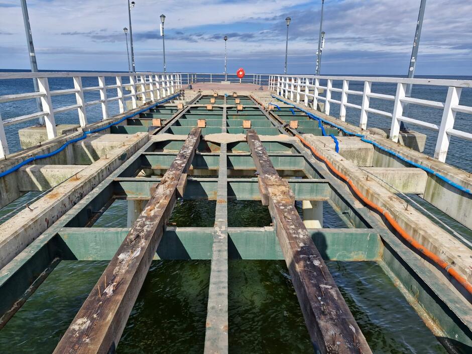 Na zdjęciu widać remontowane molo z odsłoniętą stalową konstrukcją nośną, na której częściowo ułożone są drewniane belki. Po obu stronach biegną instalacje w osłonach, a w tle rozciąga się morze i niebo z lekkim zachmurzeniem.
