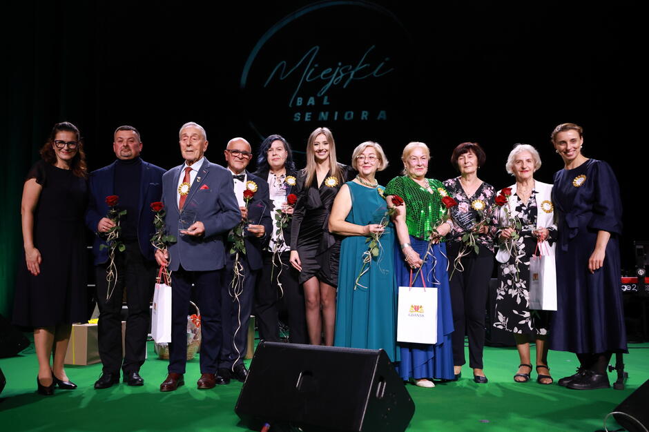 Na zdjęciu widać grupę seniorów i osób towarzyszących podczas uroczystości Miejski Bal Seniora , wszyscy uśmiechają się do kamery, trzymając róże i upominki. W tle widnieje napis wydarzenia, a cała scena ma elegancki, uroczysty charakter.

