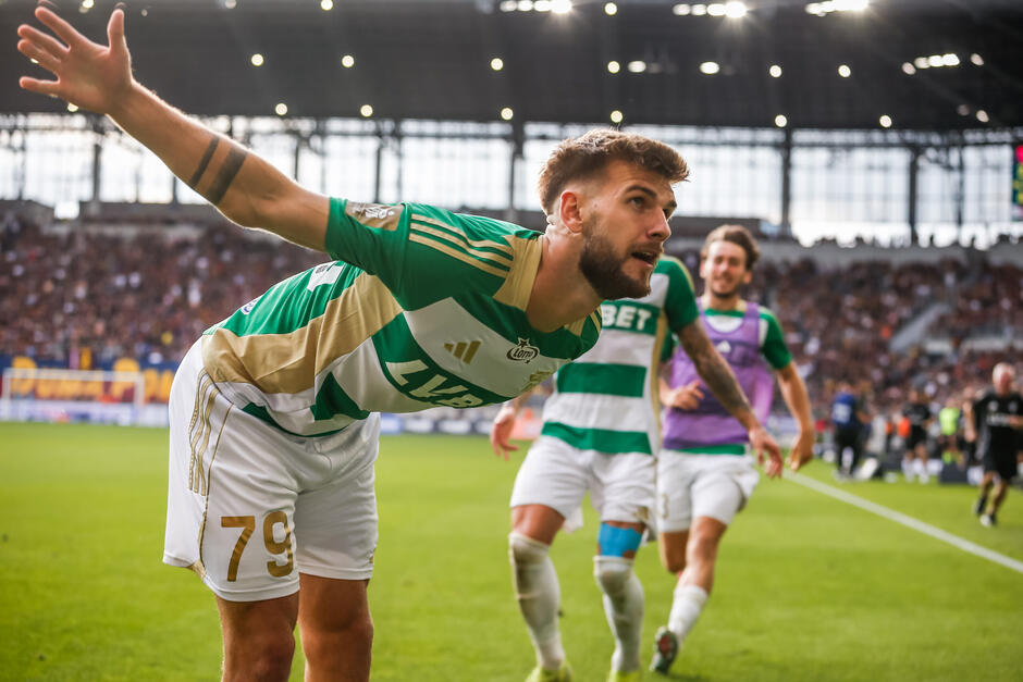 Zawodnik w biało-zielonym stroju w ukłonie. Podbiegają do niego dwaj koledzy z drużyny, z uśmiechniętymi twarzami