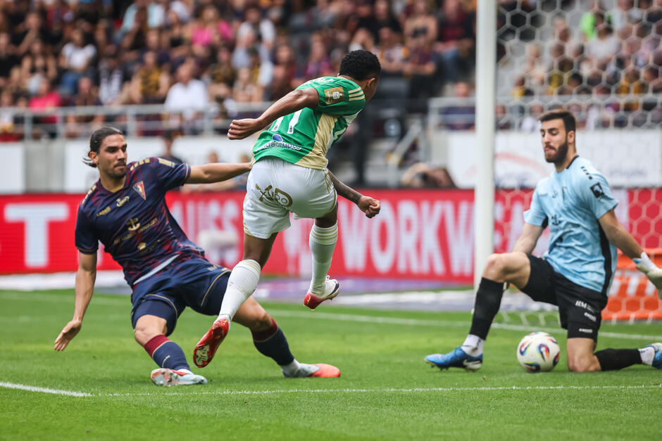 Trzej piłkarze pod bramką - jeden w biało-zielonym stroju, strzela. Drugi z lewej, w granatowym stroju, próbuje wślizgiem zablokować strzał. W głębi jest bramkarz w niskim przysiadzie, ma błękitną bluzę i czarne spodenki. Piłka jest między nogami bramkarza