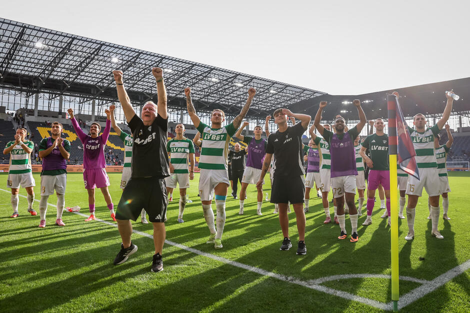 Około dwudziestu piłkarzy i członków sztabu drużyny - stoją w narożniku boiska z uniesionymi rękami i śpiewają w kierunku trybun 