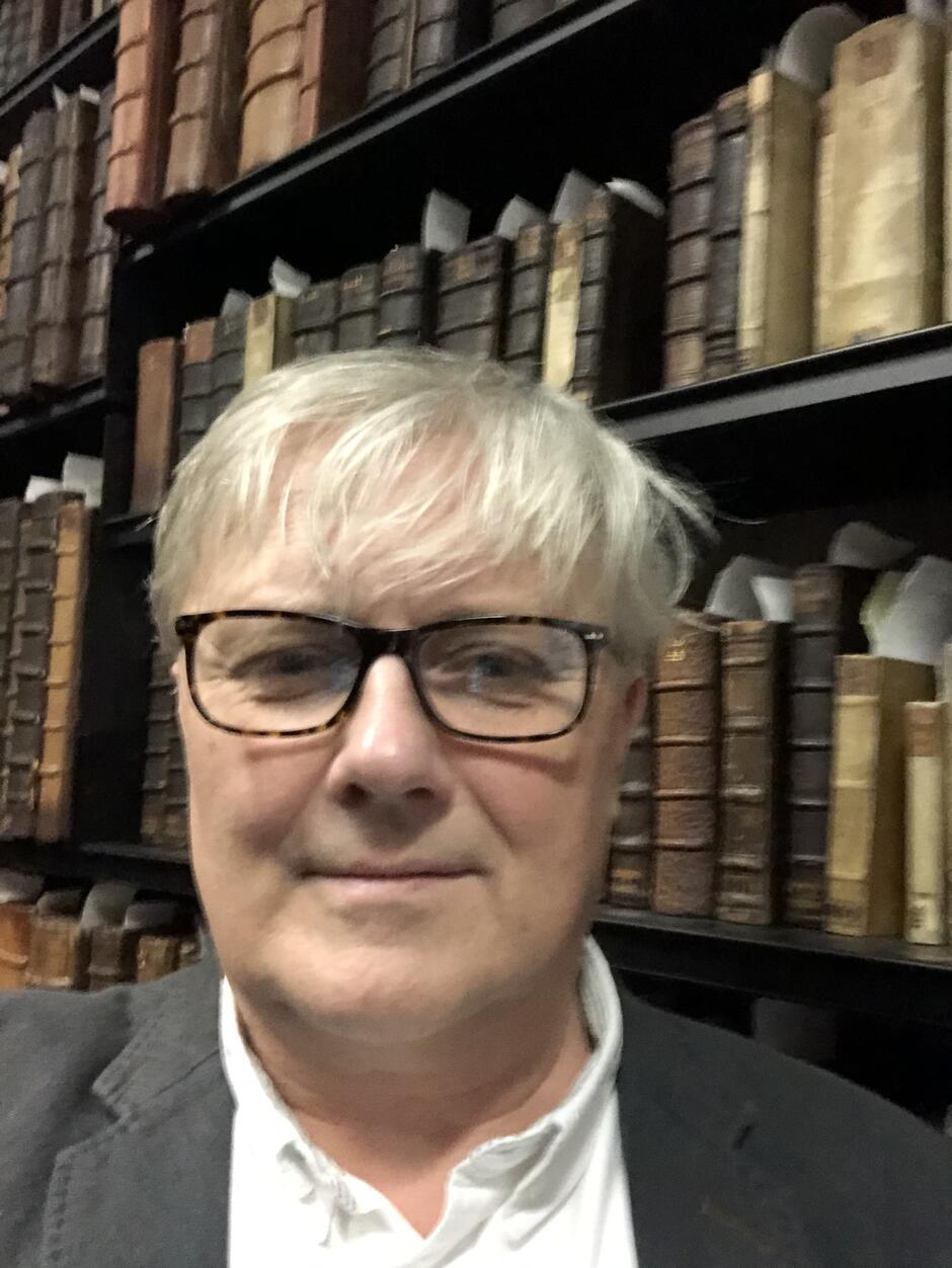 na zdjęciu mężczyzna w okularach z czarnymi oprawkami, patrzy na wprost, ma siwe włosy. Znajduje się na tle regałów z książkami.