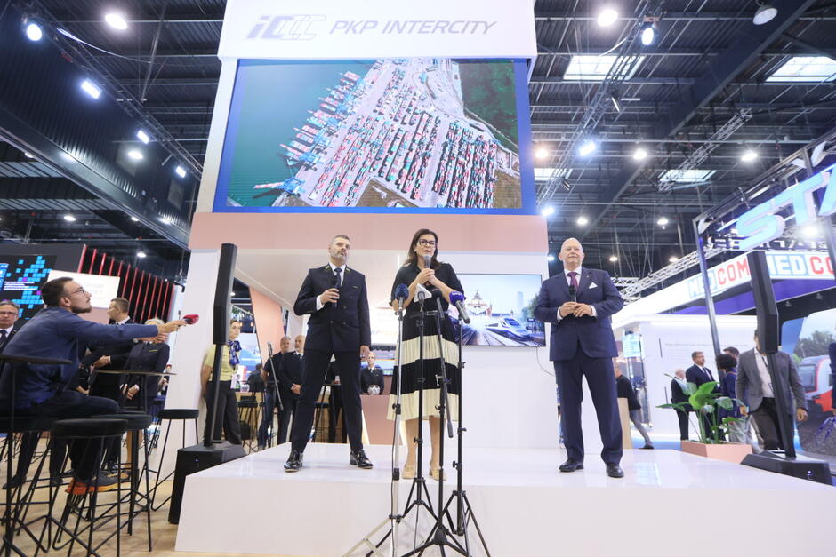 Kobieta i dwóch mężczyzn na scenie w hali, gdzie odbywają się targi. Kobieta przemawia do mikrofonu.
