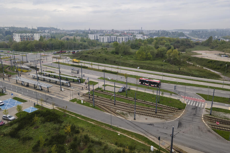 Pętla tramwajowa z kilkoma torami i peronami, obok której jedzie czerwono-biały autobus. W tle znajdują się bloki mieszkalne oraz zielone wzgórze porośnięte drzewami.