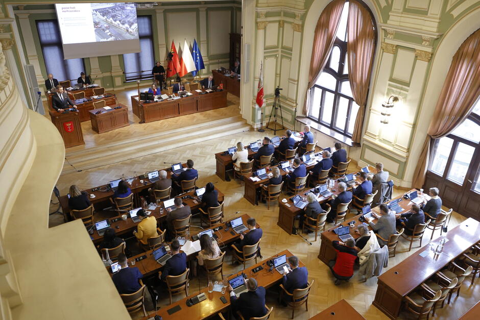 Ludzie siedzą w ławach w sali obrad rady miasta. Widok z góry na salę.