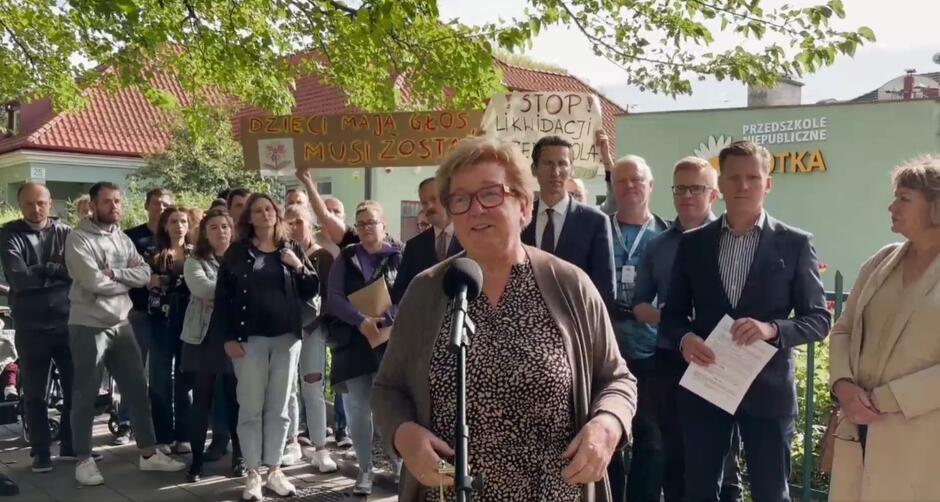 Grupa około 30 osób, przed którymi przy mikrofonie stoi kobieta w wieku ponad 60 lat 