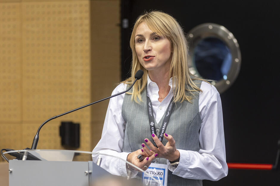 Kobieta w eleganckiej koszuli i kamizelce przemawia przy mównicy, trzymając w dłoni pilot do prezentacji. Na szyi ma identyfikator z napisem „TRAKO”, co wskazuje na udział w wydarzeniu branżowym. 