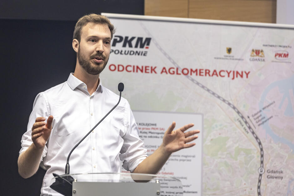 Mężczyzna w białej koszuli przemawia przy mównicy, gestykulując dłońmi. Za nim znajduje się plansza z mapą i napisem „PKM Południe – Odcinek Aglomeracyjny”. 