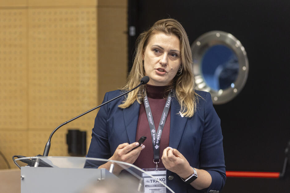 Kobieta w granatowej marynarce przemawia przy mównicy, trzymając w dłoni pilot do prezentacji. Na szyi ma identyfikator z napisem „TRAKO”, wskazujący na udział w wydarzeniu targowym. 