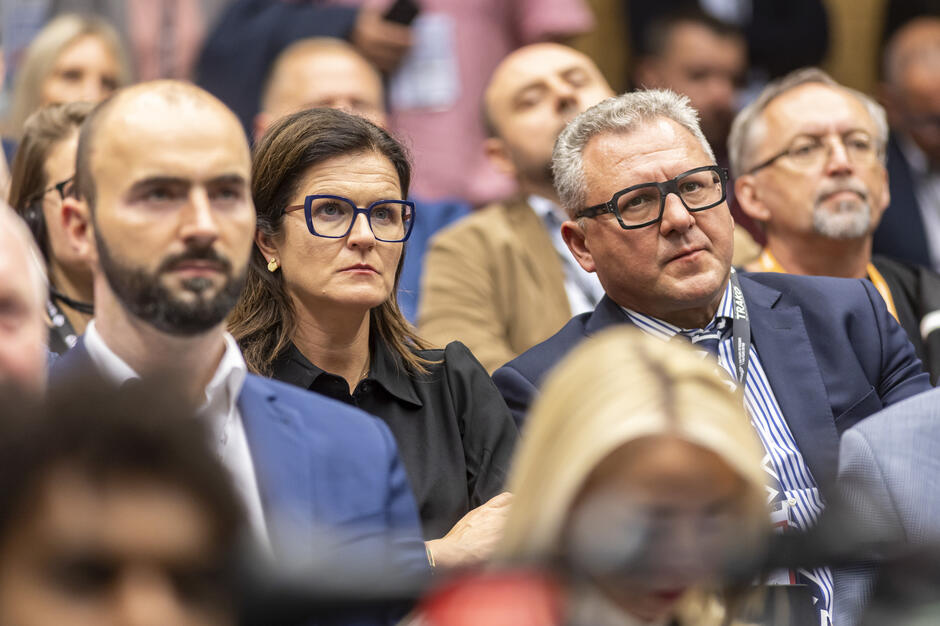 Grupa osób w eleganckich strojach siedzi na sali konferencyjnej, uważnie słuchając wystąpienia. Widać skupienie na ich twarzach, co podkreśla powagę wydarzenia. 