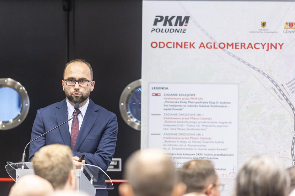 Mężczyzna w garniturze przemawia przy mównicy, trzymając pilot do prezentacji. Za nim widoczna jest plansza z mapą i napisem „PKM Południe – Odcinek Aglomeracyjny”. 
