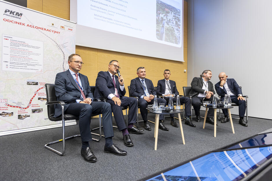 Sześciu mężczyzn w garniturach siedzi na scenie podczas panelu dyskusyjnego, a jeden z nich trzyma mikrofon i zabiera głos. Obok widoczna jest plansza z mapą „PKM Południe – Odcinek Aglomeracyjny” oraz fragment prezentacji wyświetlanej na ekranie. 