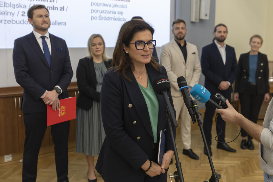 na zdjęciu kobieta w czarnej marynarce, mówi do mikrofonów dziennikarskich, za nią stoi kilka osób