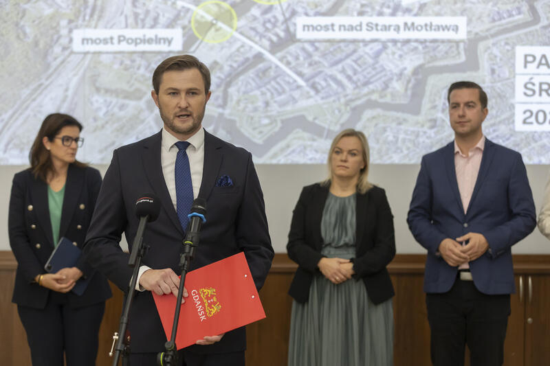 na zdjęciu mężczyzna w średnim wieku, mówi do mikrofonu, trzyma czerwoną podkładkę w ręce, za nim stoi kilka osób