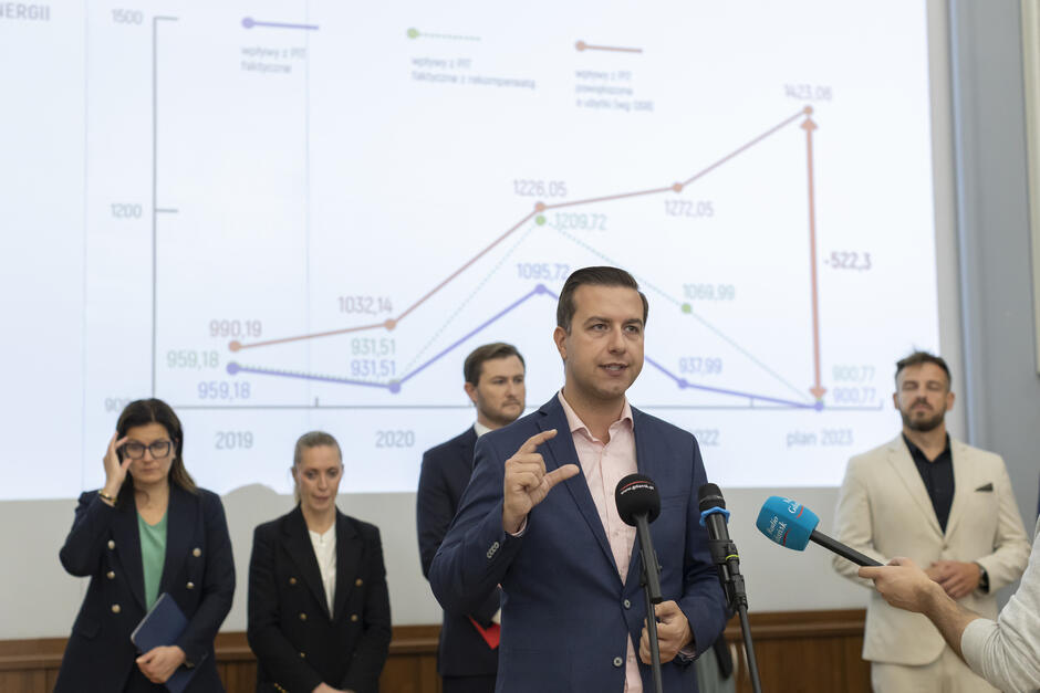 na zdjęciu mężczyzna w średnim wieku, w niebieskiej marynarce, mówi do mikrofonów dziennikarskich, za nim stoi kilka osób