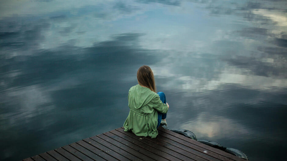 Kobieta w zielonej kurtce siedzi samotnie na drewnianym pomoście nad wodą. Wpatruje się w spokojną taflę jeziora odbijającą niebo i chmury.