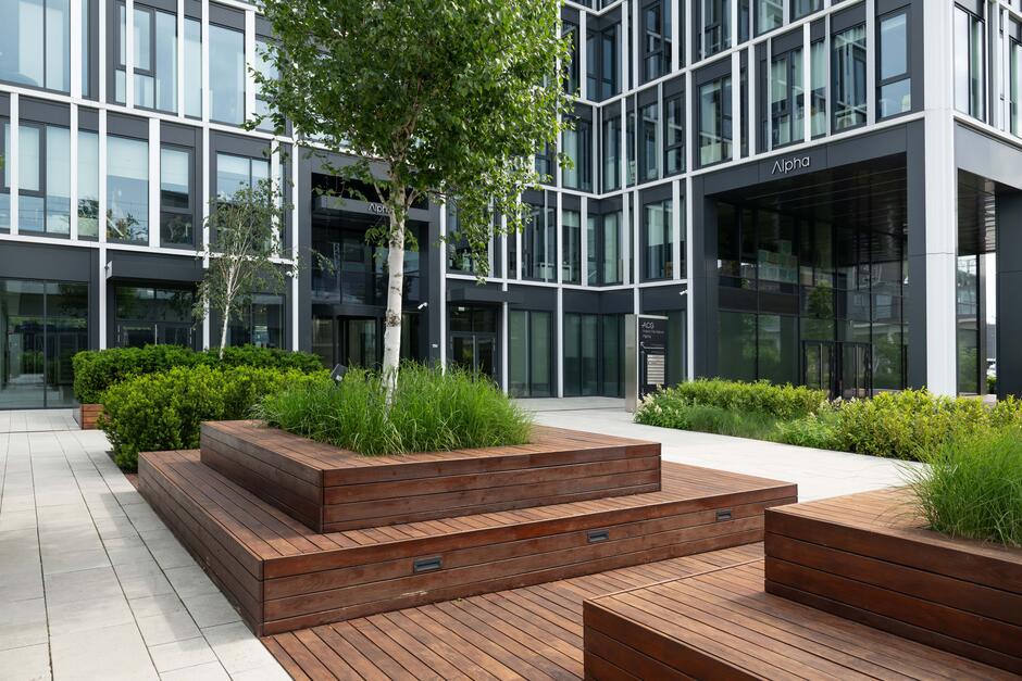 Front nowoczesnego biurowca, przed którym rośnie zieleń i drzewa.