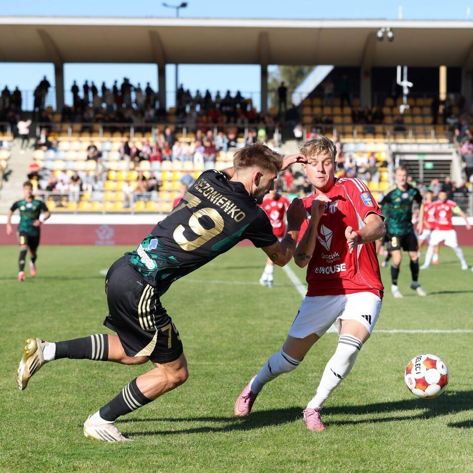 Dwaj zawodnicy walczą o piłkę na boisku. Sezonienko w czarnym kostiumie ze złotymi napisami