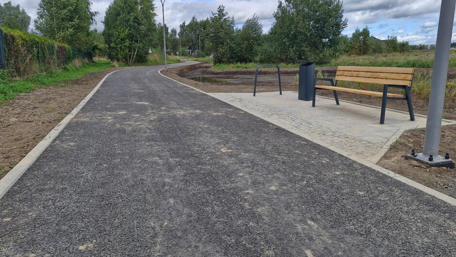 na zdjęciu po prawej ławka, kosz na śmieci i stojak rowerowy, po lewej szeroka czarna droga rowerowa