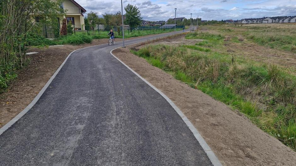 na zdjęciu nowa czarna droga rowerowa, w tle mały chłopiec na rowerze, po lewej fragment domu jednorodzinnego