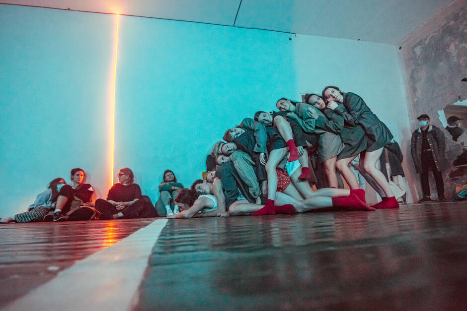 Fotografia ukazuje grupę młodych osób tańczących w klubowej przestrzeni, oświetlonej niebiesko-czerwonym światłem, z intensywną ekspresją ruchów i skupieniem na twarzach.