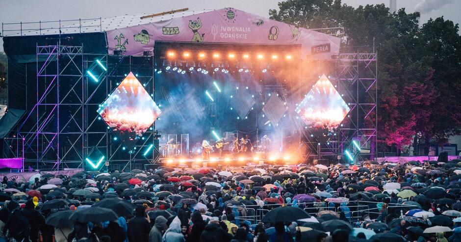 Scena plenerowa podczas Juwenaliów Gdańskich oświetlona kolorowymi reflektorami i ekranami LED, na których widać występ muzyków. Przed sceną tłum ludzi ogląda koncert, trzymając otwarte parasole w deszczu.