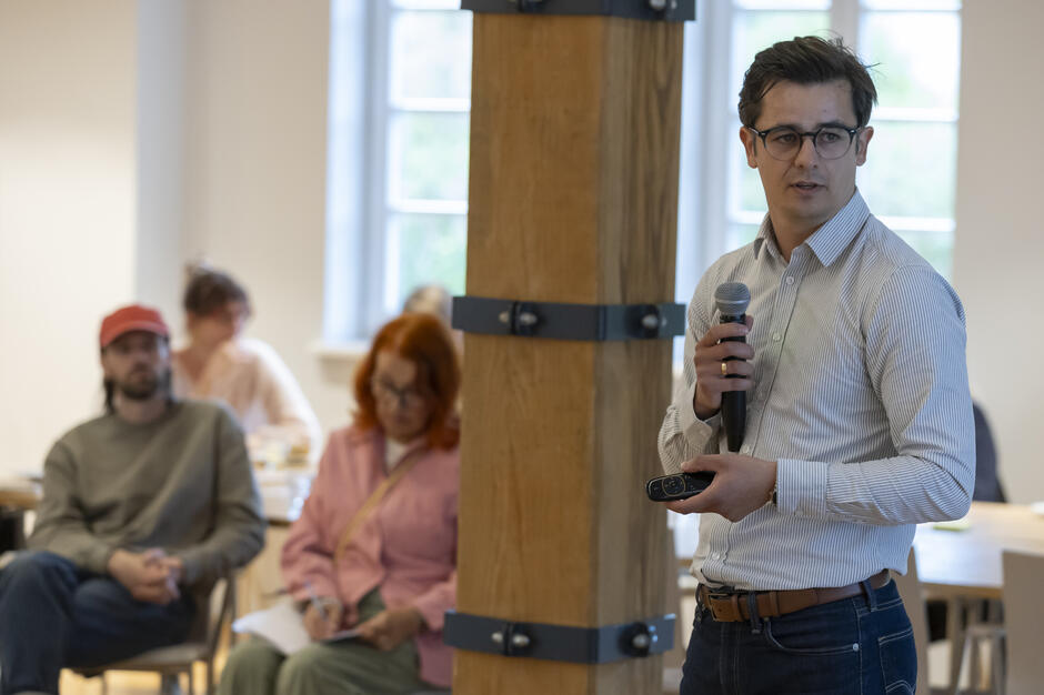 Na zdjęciu mężczyzna w okularach trzyma mikrofon i pilot do prezentacji, prowadząc wystąpienie podczas spotkania. W tle widać uczestników siedzących przy stołach, notujących i przysłuchujących się prelekcji.
