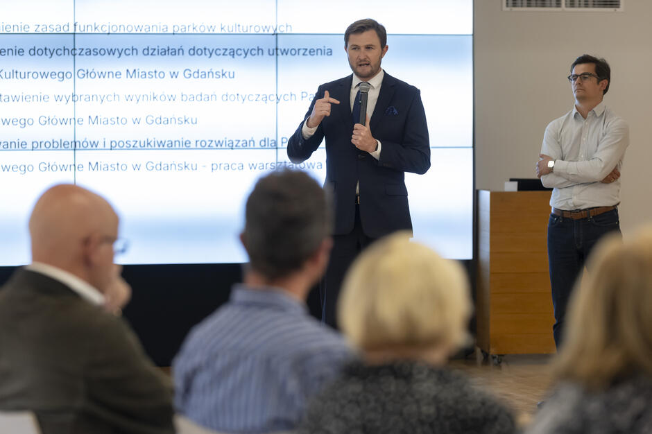 Na zdjęciu mężczyzna w garniturze przemawia do uczestników, trzymając mikrofon i gestykulując w trakcie wystąpienia. Obok niego stoi drugi prelegent, a w pierwszym planie widoczna jest publiczność słuchająca prezentacji.
