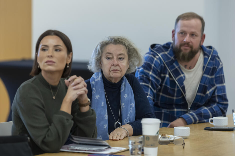 Na zdjęciu trzy osoby siedzą przy stole, uważnie przysłuchując się spotkaniu. Na blacie leżą notatki, filiżanki oraz okulary, co podkreśla roboczy charakter wydarzenia.
