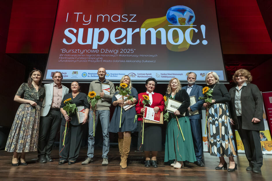 Laureaci i organizatorzy XIV Gali „Bursztynowe Dźwigi 2025” stoją wspólnie na scenie, trzymając dyplomy oraz kwiaty. W tle widoczny jest ekran z hasłem „I Ty masz supermoc!” oraz informacjami o wydarzeniu organizowanym przez Fundację Hospicyjną.
