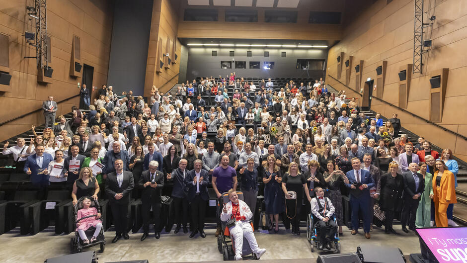 Sala wypełniona jest uczestnikami gali, którzy wspólnie pozują do pamiątkowego zdjęcia, uśmiechając się i machając w stronę aparatu. Widać różnorodność wiekową i integracyjną – wśród obecnych są zarówno dzieci, dorośli, jak i osoby z niepełnosprawnościami.

