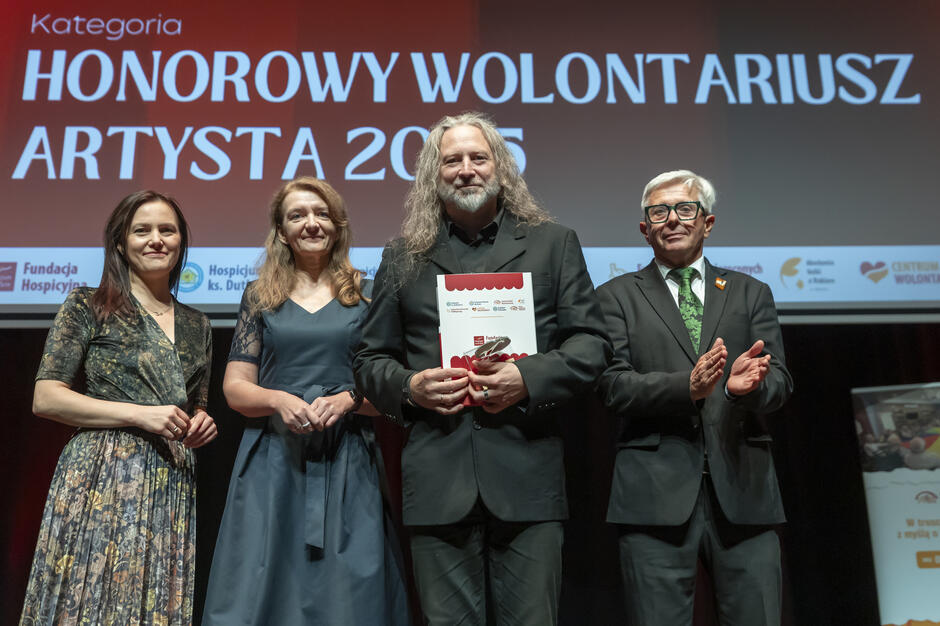 Na scenie stoi laureat nagrody „Honorowy Wolontariusz Artysta 2025” wraz z osobami wręczającymi wyróżnienie. Uśmiechy i brawa podkreślają uroczysty i pełen uznania charakter chwili.
