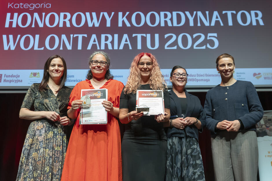 Na scenie wyróżniono laureatki nagrody „Honorowy Koordynator Wolontariatu 2025”, które dumnie prezentują swoje dyplomy. Towarzyszą im przedstawicielki organizatorów, podkreślając rangę i uroczysty charakter chwili. 