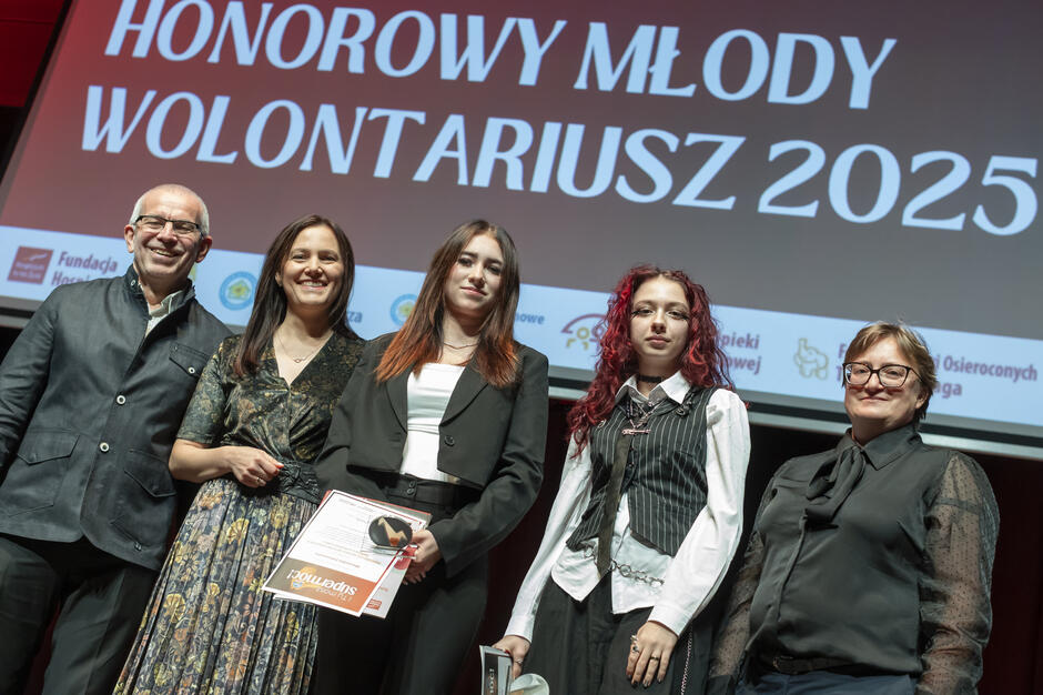 Na scenie nagrodzono laureatki tytułu „Honorowy Młody Wolontariusz 2025”, które trzymają w dłoniach dyplomy i statuetki. Towarzyszą im przedstawiciele organizatorów, podkreślając wagę zaangażowania młodych ludzi w wolontariat. 