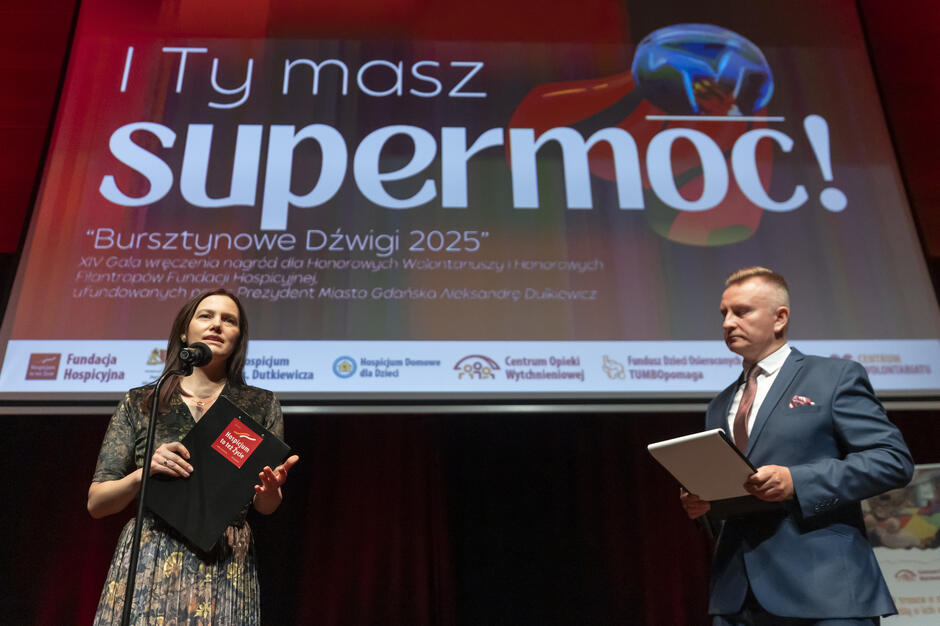 Prowadzący galę „Bursztynowe Dźwigi 2025” stoją na scenie, trzymając w dłoniach notatniki z programem wydarzenia. W tle widoczny jest duży ekran z hasłem „I Ty masz supermoc!”, które nadaje uroczystości wyjątkowego charakteru.
