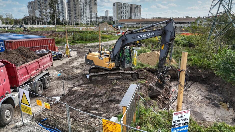 na zdjęciu żółta koparka na pierwszym planie, widać rozkopany gruntowy teren i plac budowy, który jest ogrodzony