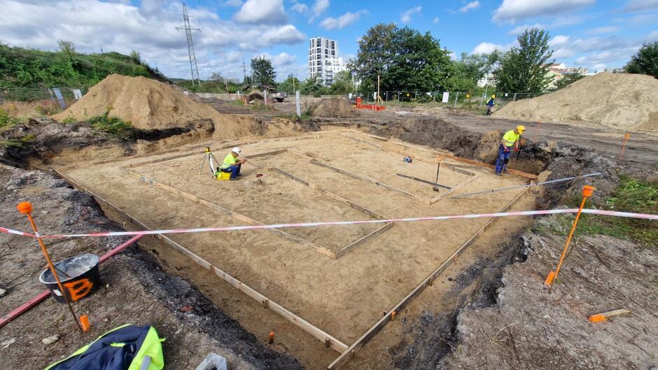 na zdjęciu duży kwadratowy plac, widać jasny piasek i dwóch mężczyzn prowadzących prace na tym terenie