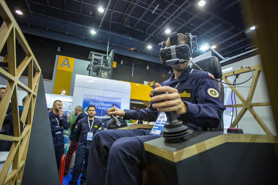 Mężczyzna w mundurze siedzi z goglami VR na głowie w hali targów.