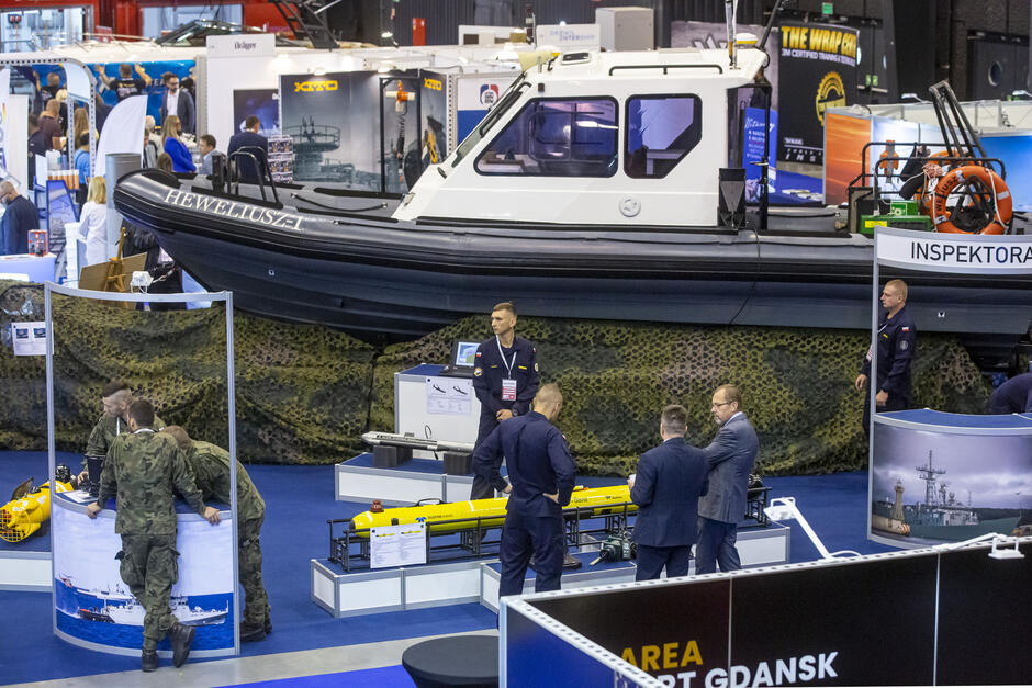 Zdjęcie z wyższego poziomu, widok na halę, gdzie odbywają się targi. Na stoisku widać motorówkę oraz ludzi wokół.