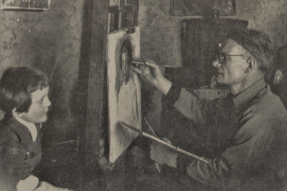 Czarno-biała fotografia przedstawia malarza pracującego nad portretem siedzącego naprzeciwko dziecka. Artysta skupiony na pracy trzyma pędzel i maluje twarz modela na sztaludze. 