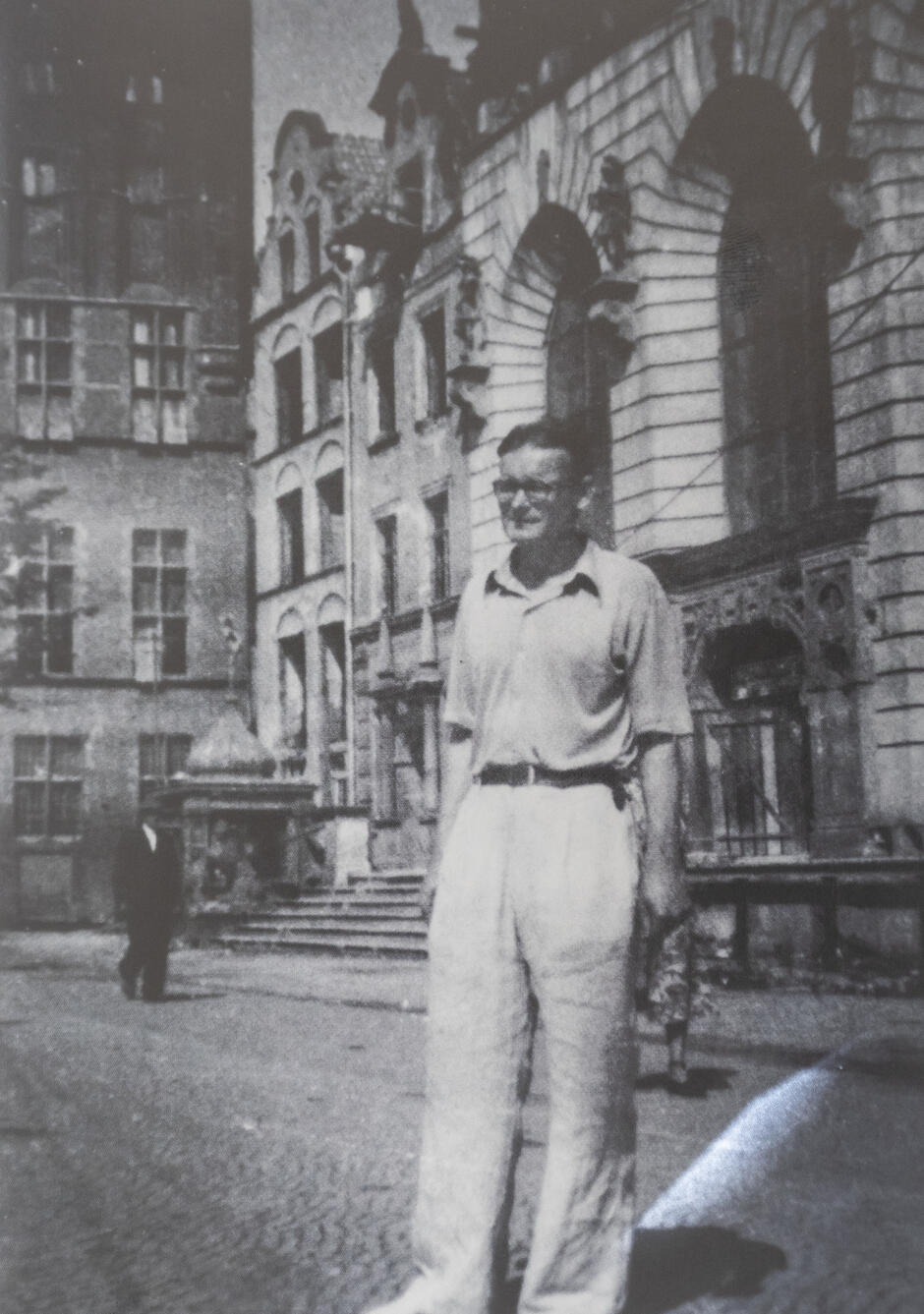 Czarno-biała fotografia przedstawia mężczyznę stojącego na tle kamienic i zabytkowych budynków w Gdańsku. Ubrany w jasne spodnie i koszulę z krótkim rękawem, patrzy w stronę aparatu w pogodny dzień. 