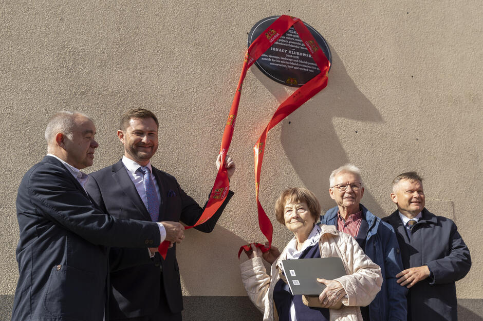Grupa osób odsłania pamiątkową tablicę poświęconą Ignacemu Klukowskiemu, trzymając czerwoną wstęgę z herbem. Uroczystość odbywa się przy słonecznej pogodzie, a uczestnicy stoją uśmiechnięci przed ścianą budynku.
