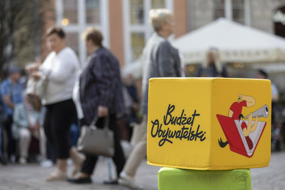 na zdjęciu dwie duże pluszowe kostki, jedna żółta z czarnym napisem budżet obywatelski, w tle przechodzą trzy kobiety
