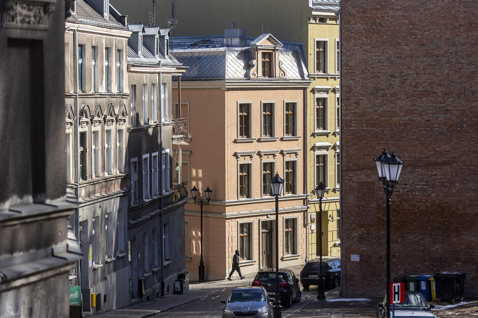 Wąska brukowana uliczka otoczona jest kamienicami o odnowionych fasadach w pastelowych kolorach, na których widać ozdobne detale architektoniczne. Na rogu różowej kamienicy idzie pieszy, a wzdłuż ulicy stoją zaparkowane samochody i klasyczne latarnie.
