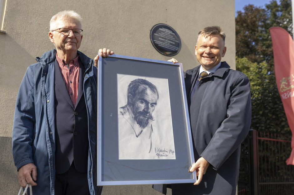 Dwaj mężczyźni stoją obok siebie, trzymając w ramie rysunek portretowy brodatego mężczyzny. W tle widoczna jest tablica pamiątkowa umieszczona na ścianie budynku oraz fragment czerwonego sztandaru.
