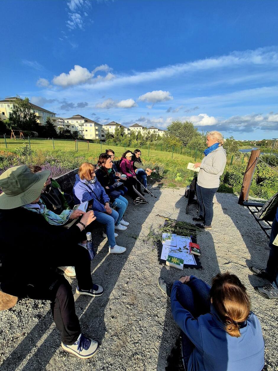 Grupa osób uczestniczy w spotkaniu edukacyjnym na świeżym powietrzu, gdzie prowadząca opowiada o roślinach i pokazuje materiały dydaktyczne. Uczestnicy siedzą na ławkach i słuchają, a przed nimi na ziemi rozłożone są książki i zebrane okazy roślin.
