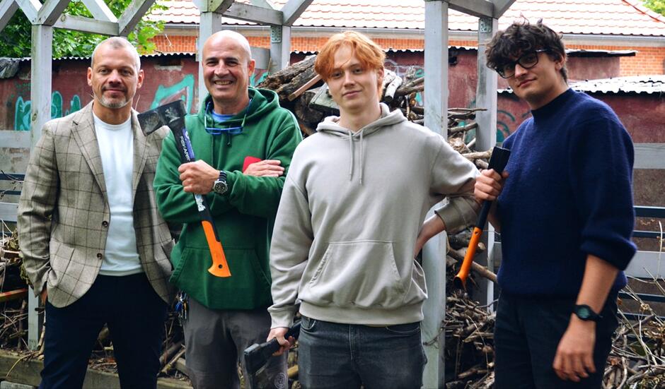 Czterech mężczyzn na tle altanki z drewnem, trzech trzyma siekiery